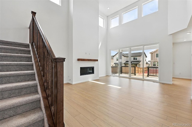 unfurnished living room featuring stairway, a glass covered fireplace, a towering ceiling, light wood finished floors, and baseboards
