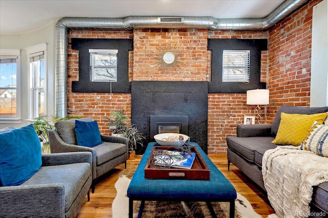 living area with a glass covered fireplace, wood finished floors, visible vents, and brick wall