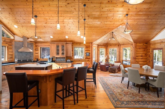 kitchen with log walls, lofted ceiling, a kitchen island with sink, wood ceiling, and wall chimney range hood