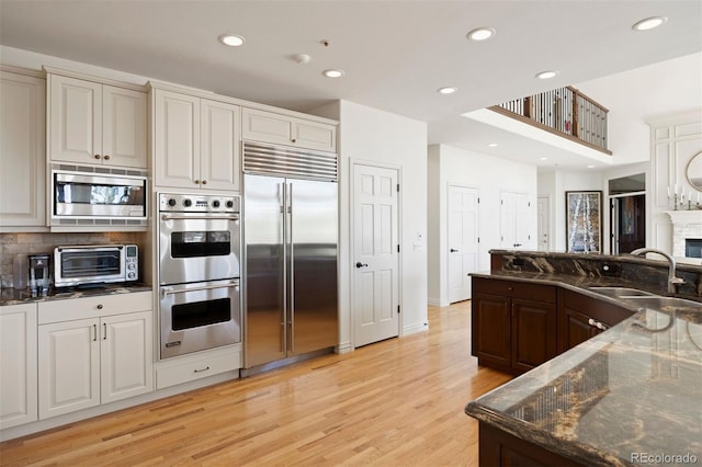 kitchen with built in appliances, light wood-style flooring, a sink, dark stone counters, and a glass covered fireplace