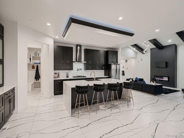 kitchen with appliances with stainless steel finishes, backsplash, a kitchen bar, a center island with sink, and wall chimney range hood