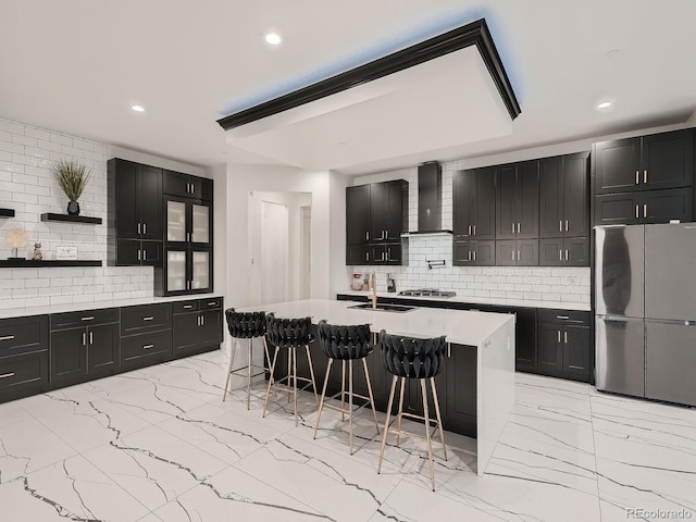 kitchen featuring decorative backsplash, wall chimney exhaust hood, a breakfast bar area, stainless steel appliances, and a kitchen island with sink