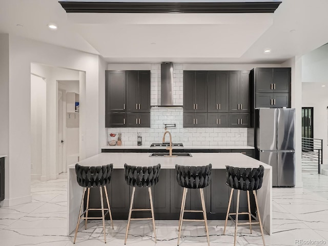 kitchen with stainless steel fridge, tasteful backsplash, a kitchen island with sink, wall chimney range hood, and a breakfast bar area