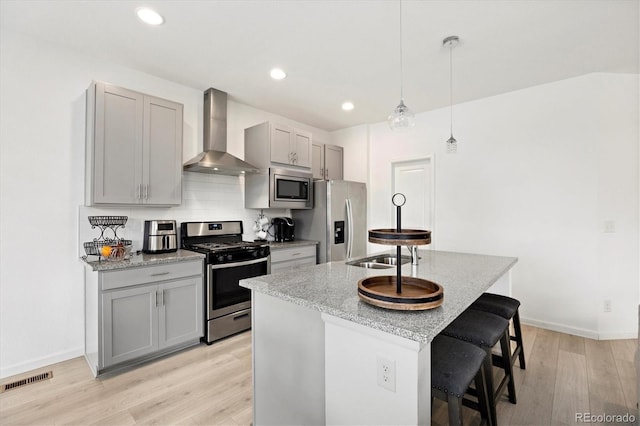 kitchen with stainless steel appliances, wall chimney range hood, an island with sink, light hardwood / wood-style floors, and gray cabinets