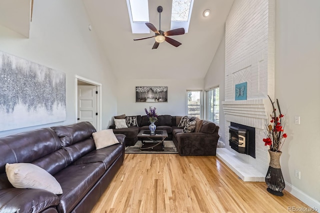 living area featuring high vaulted ceiling, a skylight, ceiling fan, light wood-style flooring, and a brick fireplace