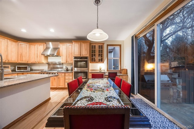 kitchen with light wood finished floors, light brown cabinetry, double oven, wall chimney range hood, and decorative light fixtures