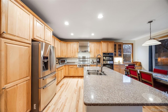 kitchen with light brown cabinets, wall chimney range hood, an island with sink, appliances with stainless steel finishes, and a sink