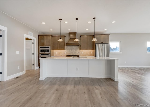 kitchen featuring tasteful backsplash, decorative light fixtures, an island with sink, custom range hood, and stainless steel appliances