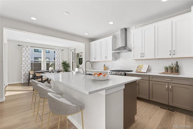 kitchen with a kitchen island with sink, light hardwood / wood-style floors, a breakfast bar, wall chimney exhaust hood, and white cabinets