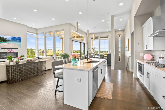 kitchen featuring pendant lighting, sink, wall chimney exhaust hood, white cabinetry, and a kitchen island with sink