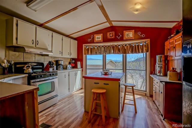 kitchen with lofted ceiling, stainless steel gas range oven, under cabinet range hood, backsplash, and light wood-style floors