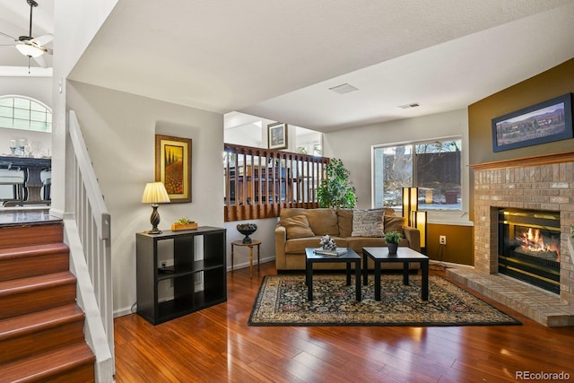 living room with a brick fireplace, dark wood-type flooring, and ceiling fan