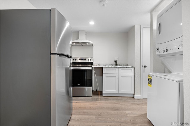 kitchen with light wood finished floors, stacked washer / drying machine, appliances with stainless steel finishes, a sink, and wall chimney exhaust hood