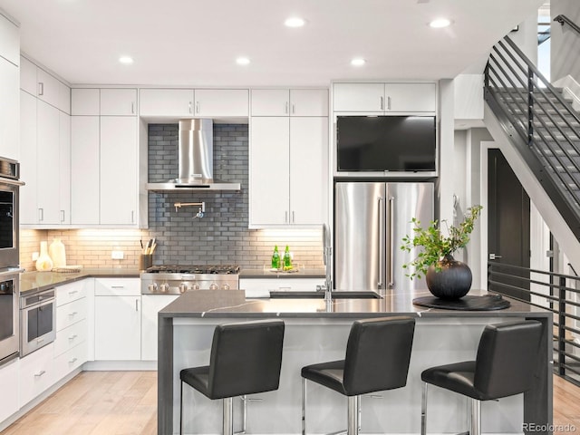 kitchen with appliances with stainless steel finishes, white cabinets, a kitchen breakfast bar, and wall chimney exhaust hood