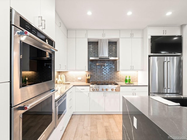 kitchen featuring stone countertops, wall chimney range hood, stainless steel appliances, decorative backsplash, and white cabinets