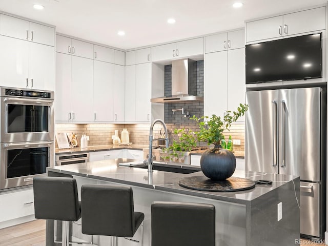 kitchen with light wood finished floors, tasteful backsplash, wall chimney exhaust hood, appliances with stainless steel finishes, and a kitchen bar