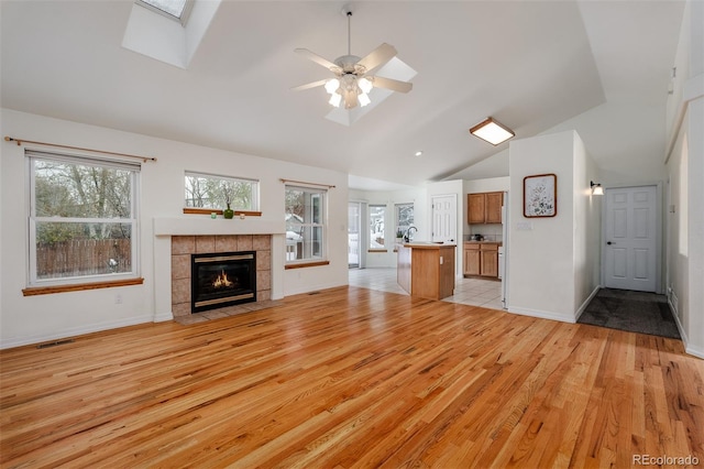 unfurnished living room featuring light wood-type flooring, ceiling fan, vaulted ceiling with skylight, and a tiled fireplace