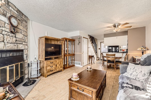 living room with a textured ceiling, ceiling fan, light hardwood / wood-style floors, and a stone fireplace