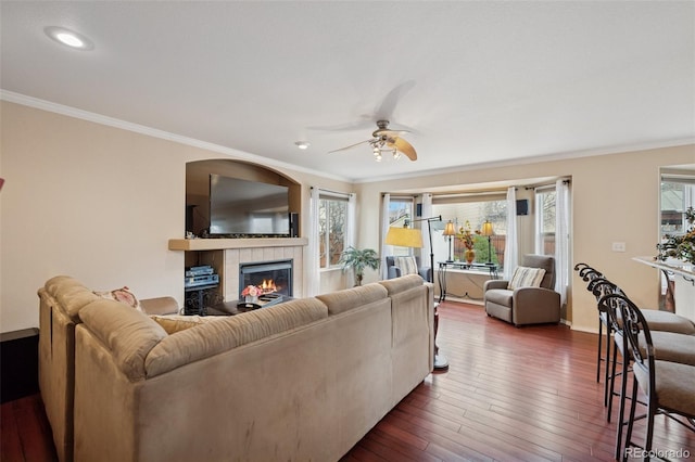 living room featuring dark wood-style flooring, baseboards, a fireplace, ornamental molding, and a ceiling fan