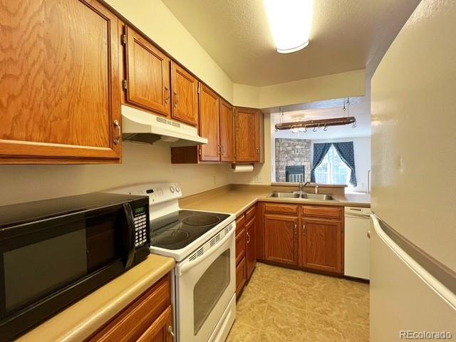 kitchen with white appliances, brown cabinetry, under cabinet range hood, light countertops, and a sink