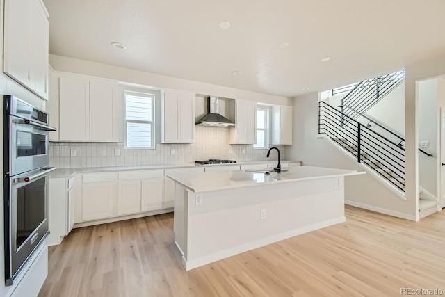 kitchen featuring sink, wall chimney range hood, a kitchen island with sink, white cabinets, and light wood-type flooring