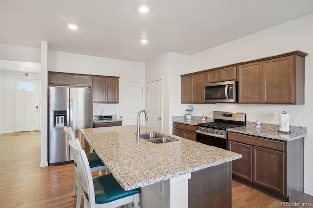 kitchen featuring light wood-type flooring, light stone counters, stainless steel appliances, a kitchen island with sink, and sink