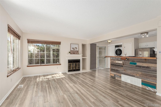 unfurnished living room featuring stacked washer / dryer, a textured ceiling, baseboards, light wood finished floors, and a fireplace with flush hearth