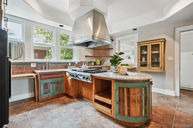 kitchen with baseboards, glass insert cabinets, island exhaust hood, stainless steel gas cooktop, and a sink