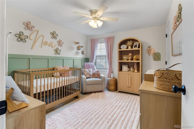 bedroom with light colored carpet, a decorative wall, a ceiling fan, wainscoting, and a crib