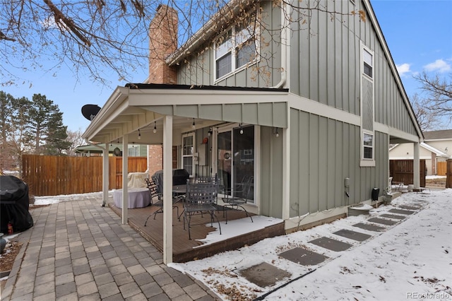 snow covered property featuring a patio, board and batten siding, a chimney, and fence