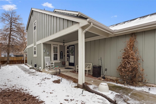 snow covered rear of property featuring board and batten siding, fence, and a porch