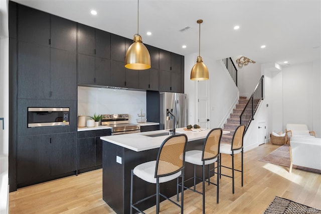 kitchen with stainless steel appliances, a sink, a center island with sink, and dark cabinets