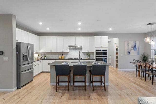 kitchen with decorative light fixtures, white cabinetry, stainless steel appliances, an island with sink, and light wood-type flooring