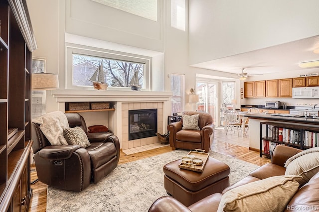 living room featuring a tiled fireplace, ceiling fan, light wood-type flooring, and a towering ceiling