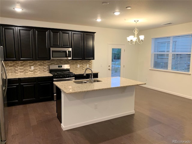 kitchen featuring a chandelier, stainless steel appliances, light stone counters, dark wood-type flooring, and a center island with sink