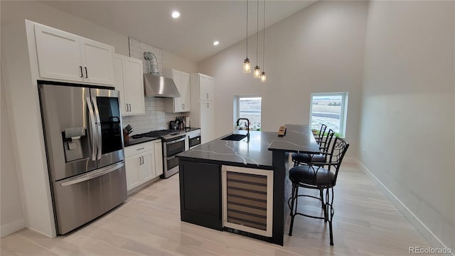 kitchen with wall chimney range hood, white cabinets, an island with sink, stainless steel appliances, and decorative backsplash