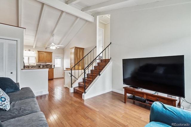 living area featuring stairway, light wood finished floors, visible vents, a ceiling fan, and vaulted ceiling with beams