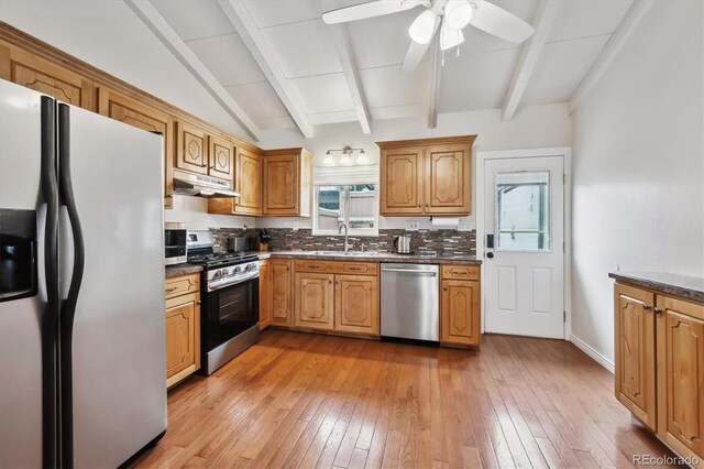 kitchen featuring a sink, lofted ceiling with beams, under cabinet range hood, stainless steel appliances, and dark countertops