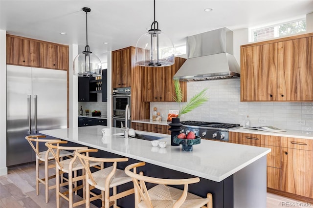 kitchen with wall chimney exhaust hood, a kitchen island with sink, stainless steel appliances, and light hardwood / wood-style floors