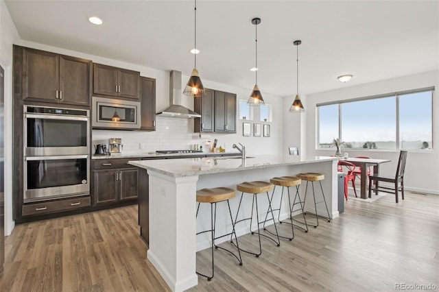 kitchen with dark brown cabinetry, appliances with stainless steel finishes, light wood finished floors, a kitchen breakfast bar, and wall chimney exhaust hood