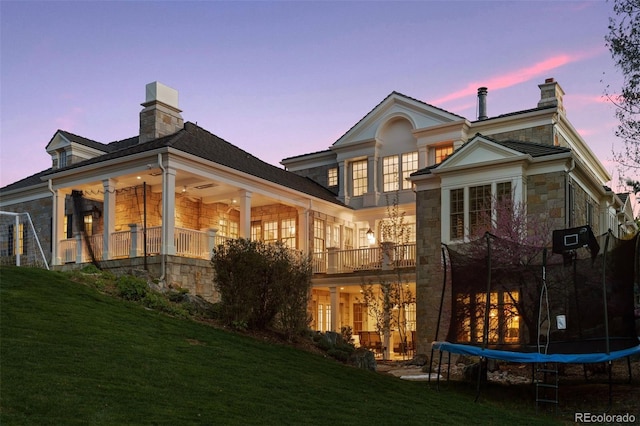 back house at dusk with a balcony, a trampoline, and a yard