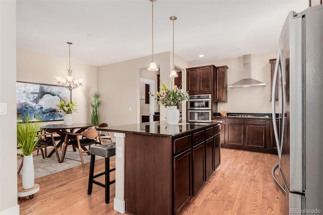 kitchen featuring appliances with stainless steel finishes, a center island, wall chimney exhaust hood, a kitchen bar, and decorative light fixtures