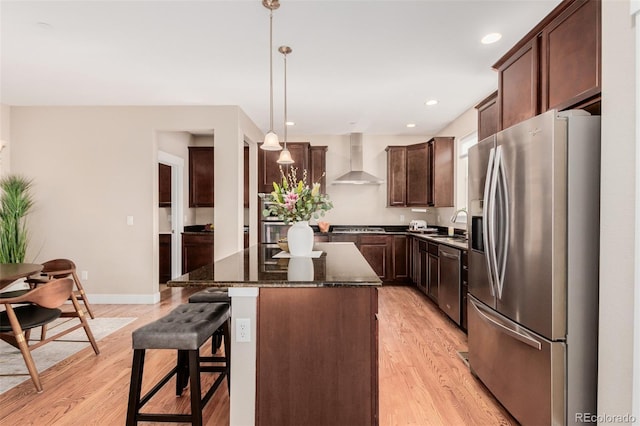 kitchen featuring hanging light fixtures, appliances with stainless steel finishes, wall chimney exhaust hood, a center island, and a breakfast bar