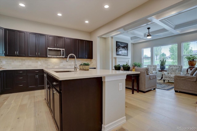 kitchen with sink, coffered ceiling, tasteful backsplash, an island with sink, and light wood-type flooring