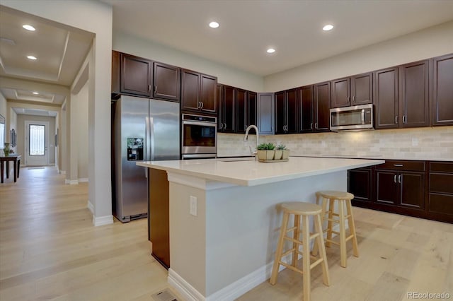 kitchen with a breakfast bar area, backsplash, stainless steel appliances, light hardwood / wood-style floors, and an island with sink