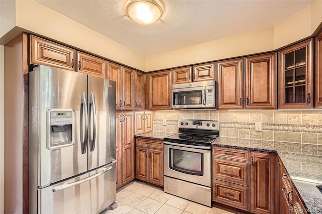 kitchen featuring dark stone counters, tasteful backsplash, brown cabinets, glass insert cabinets, and appliances with stainless steel finishes