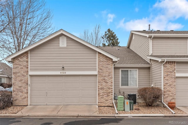 view of front facade with central AC unit, an attached garage, brick siding, a shingled roof, and driveway