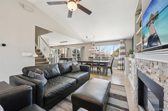 living area with light tile patterned flooring, a stone fireplace, ceiling fan with notable chandelier, and stairway