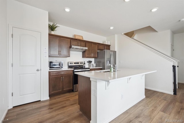608 N Bersshine Ct, Aurora, CO 80137 - kitchen featuring appliances with stainless steel finishes, under cabinet range hood, a kitchen island with sink, light countertops, and wood finished floors