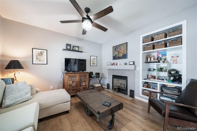 living room featuring wood-type flooring, built in features, ceiling fan, and a tile fireplace